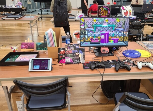 Nice Gear Games booth with a monitor and Nice Disc on the right, and a box of zines and postcards for Indie Tsushin on the left